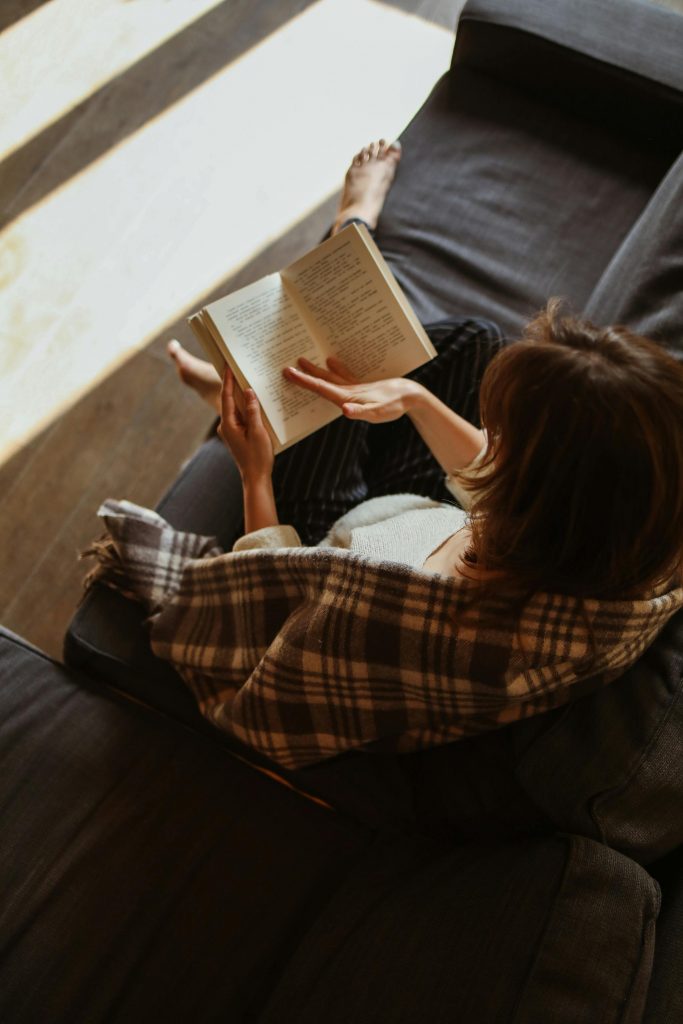 Mulher relaxando em poltrona de leitura com livro, próximo à janela com luz natural - exemplo de cantinho de leitura acolhedor